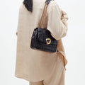 Black quilted calf leather handbag with gold buckle and chain strap, worn by a person in beige linen.