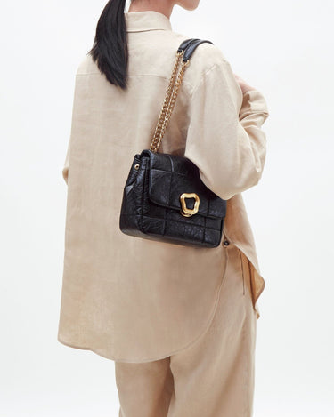 Black quilted calf leather handbag with gold buckle and chain strap, worn by a person in beige linen.