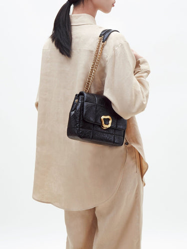 Black quilted calf leather handbag with gold buckle and chain strap, worn by a person in beige linen.