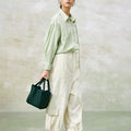 Medium Song Bag in emerald green leather, held by a woman wearing a casual green shirt and off-white cargo pants.