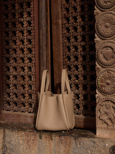 Medium Song Bag in taupe full-grain leather with dual handles against carved wood panels.