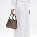 Medium Song Bag in taupe full-grain leather with textured finish and rounded handles, worn with white linen attire.