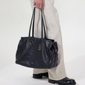 Onyx Medium Yore Duffle Bag: Black leather tote with white stitching, gold hardware. Held by person in light trousers.