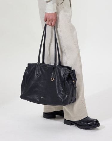 Onyx Medium Yore Duffle Bag: Black leather tote with white stitching, gold hardware. Held by person in light trousers.