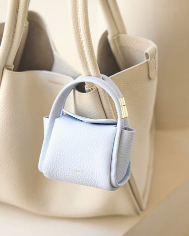 Sky Mini Song AirPods Case in textured leather with gold hardware, attached to a beige tote bag.