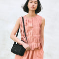 Mini Song Bag in Onyx black pebbled leather with gold hardware, worn by a woman in a peach pleated dress.