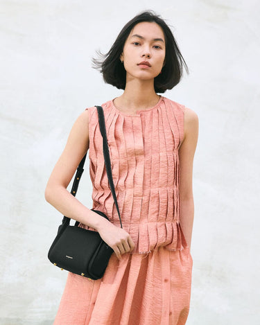Mini Song Bag in Onyx black pebbled leather with gold hardware, worn by a woman in a peach pleated dress.