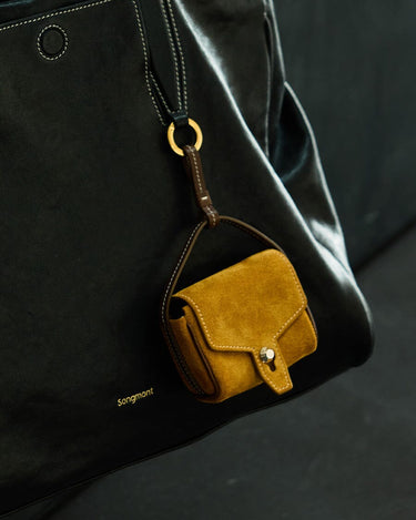 Ochre suede Mini Yore AirPods Case with snap closure, attached to a black leather Songmont handbag.