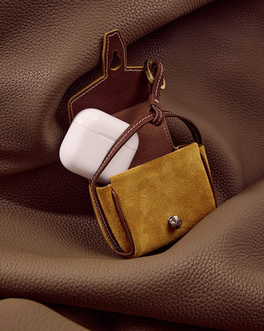 Ochre Mini Yore AirPods Case with white AirPods inside, made of suede and leather with metal clasp.