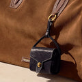 Mini Yore AirPods case in navy leather with gold clasp, attached to a tan suede tote bag.