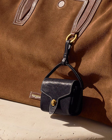 Mini Yore AirPods case in navy leather with gold clasp, attached to a tan suede tote bag.