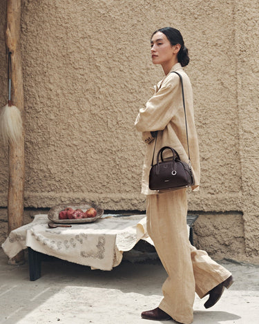 Mini Yore Bowling Bag in dark brown leather with silver hardware and white stitching, worn by a model in a beige suit.