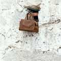Sandal brown Mini Yore Duffle Bag, vegetable-tanned leather, detailed stitching, gold hardware, on stone wall.