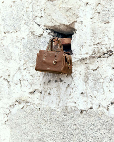 Sandal brown Mini Yore Duffle Bag, vegetable-tanned leather, detailed stitching, gold hardware, on stone wall.