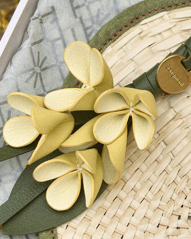 Osmanthus Charm: delicate yellow leather flower accessory with green strap and "Songmont" logo on woven straw background.