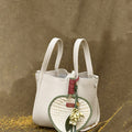 Cream textured leather handbag with heart-shaped woven straw charm, green leaves, and floral accents.