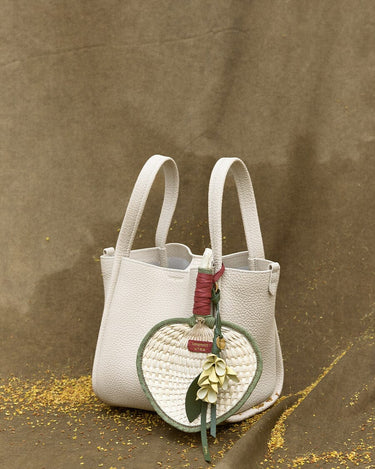 Cream textured leather handbag with heart-shaped woven straw charm, green leaves, and floral accents.