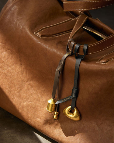 Brown leather bag with gold prayer wheel and cylindrical charms, featuring "Songmont" embossed on leather ties.