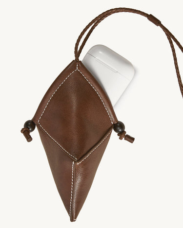 Brown leather Prism Airpods case with white stitching, braided strap, and wooden bead closure holding white AirPods case.