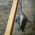 Black leather Prism Airpods Case with braided strap and sandalwood beads, hanging against a brick wall.