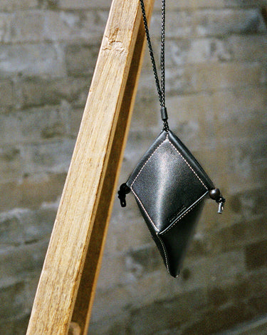 Black leather Prism Airpods Case with braided strap and sandalwood beads, hanging against a brick wall.
