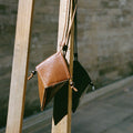 Prism Airpods Case in Sandal leather, featuring a geometric design and braided strap, hanging from a wooden structure.
