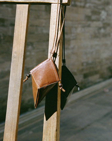 Prism Airpods Case in Sandal leather, featuring a geometric design and braided strap, hanging from a wooden structure.