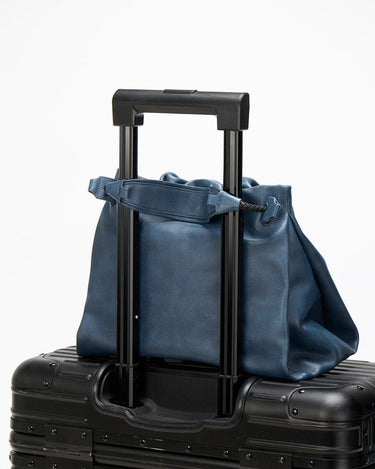 Small Gather Bag Set in navy blue leather, a versatile tote for travel, secured on a suitcase handle.