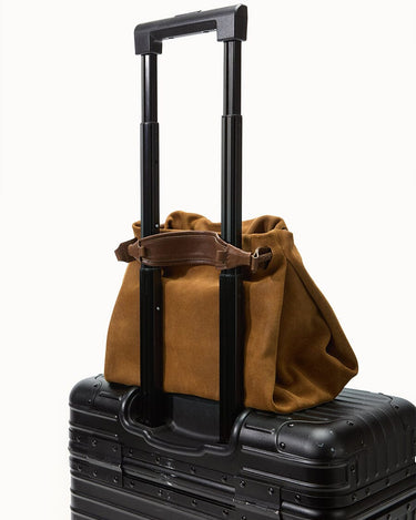 Ochre suede Small Gather Bag Set resting on a black rolling suitcase, featuring leather straps and travel-ready design.