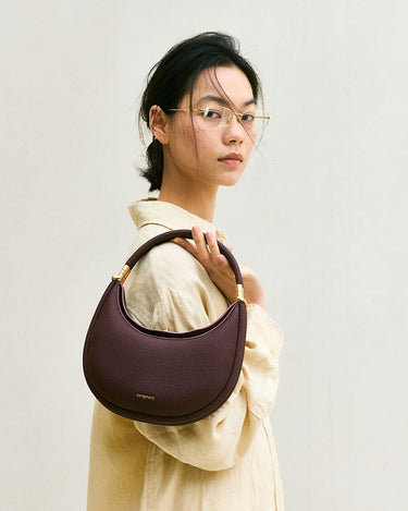 Small Luna Bag in burgundy full-grain leather with gold hardware, held by a woman wearing glasses.
