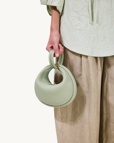 Sage green pebble-grain Small Luna Bag with twisted top handle and gold hardware, paired with a textured light green blouse and beige pants.