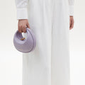 Lilac textured leather Small Luna Bag with gold hardware held by a hand, paired with a white linen shirt and wide-leg pants.