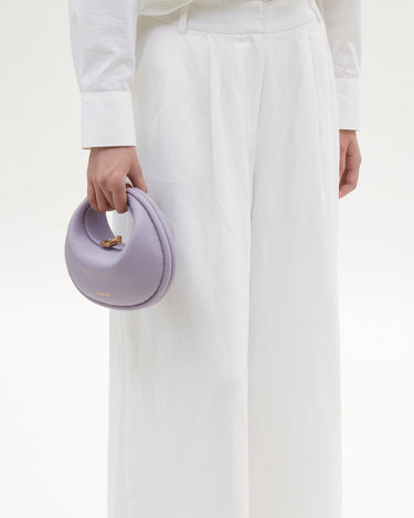 Lilac textured leather Small Luna Bag with gold hardware held by a hand, paired with a white linen shirt and wide-leg pants.