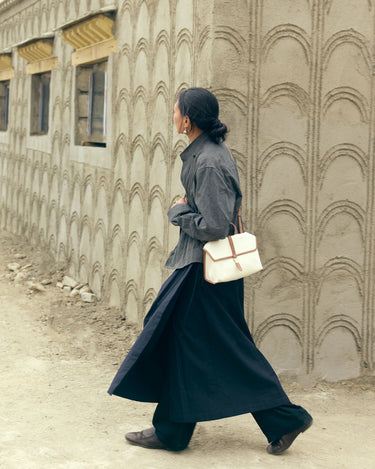 Small Shan Satchel Bag in cream canvas with tan leather trim, worn by a woman in a grey shirt and black pants walking by a textured wall.