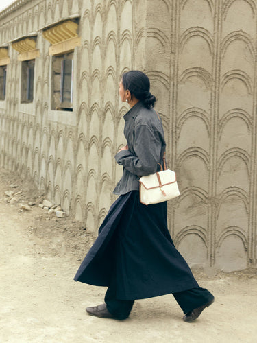 Small Shan Satchel Bag in cream canvas with tan leather trim, worn by a woman in a grey shirt and black pants walking by a textured wall.