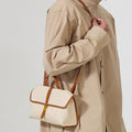 Small Shan Satchel Bag in canvas with tan leather trim and gold buckle, worn by a person in a beige trench coat.