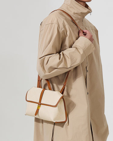 Small Shan Satchel Bag in canvas with tan leather trim and gold buckle, worn by a person in a beige trench coat.