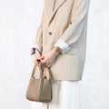 A person holds a small taupe pebble-grain leather Songmont bag, wearing a cream blazer and white pants.