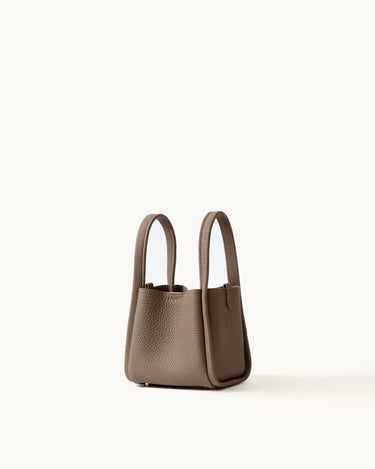 Small Song Bag in taupe pebbled full-grain leather, minimalist bucket design with dual top handles.