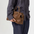 Brown Small Yore Duffle Bag in Sandal leather, white stitching, gold hardware. Carried by person in grey shirt.