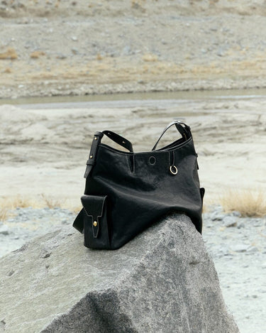 Small Yore Hobo Bag in Onyx leather, featuring adjustable strap and exterior pocket, rests on a rock outdoors.