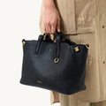 A person holds a black pebbled leather Small Yore Tote Bag by Songmont with gold hardware.