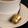 Golden cup-shaped Worry-Free Cup Charm with leather strap on speckled saucer, featuring embossed 'G' logo.