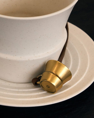 Golden cup-shaped Worry-Free Cup Charm with leather strap on speckled saucer, featuring embossed 'G' logo.