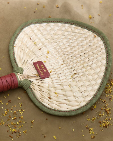 Woven fan charm made of palm fronds with dark green trim and leather handle. Features 'Songmont' tag.