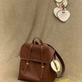 Woven fan charm on brown leather Songmont backpack with intricate stitching and gold buckles.