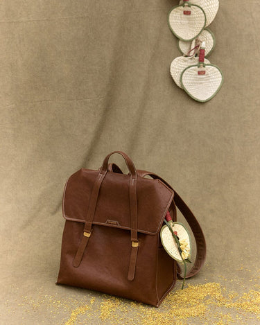 Woven fan charm on brown leather Songmont backpack with intricate stitching and gold buckles.