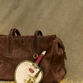 Woven fan charm with floral detail on brown suede tote bag. Songmont accessory.