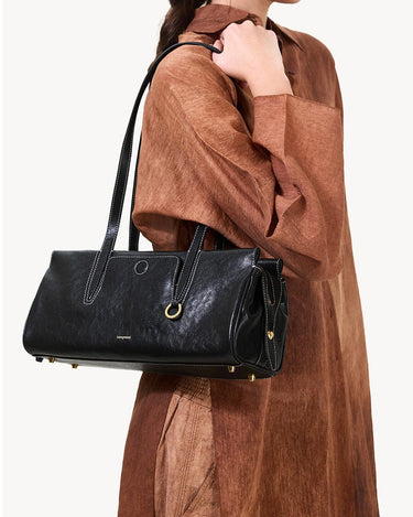 Black Yore Baguette Bag in Onyx with gold hardware and white stitching, worn by a woman in a brown dress.