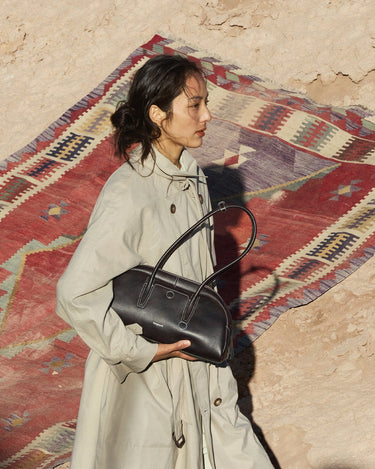 Yore Toast Bag in Ebony Nappa leather with white stitching, held by a woman in a trench coat.